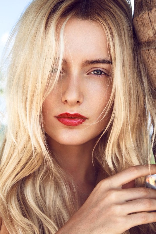 Close-up portrait of beautiful young woman with curly long blonde hair, green mysterious eyes and red sexy big lips posing next to the palm and looking to you through lens.